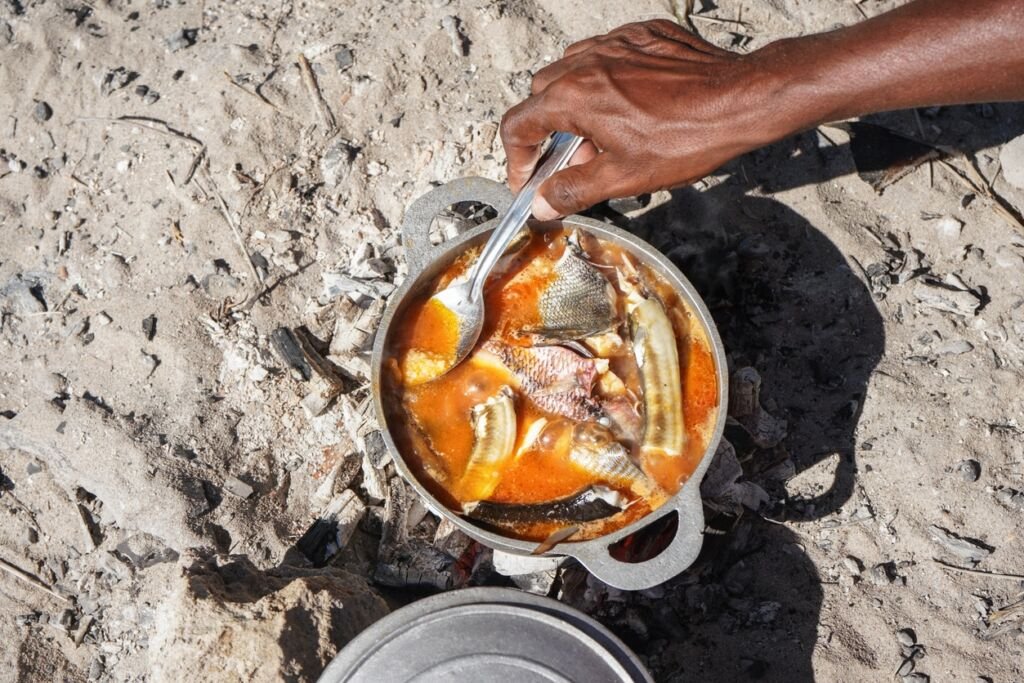 Fish soup or stew cooked from freshly catch seafood in aluminium pot, closeup detail to local man hand from above, sun shines on sand near.