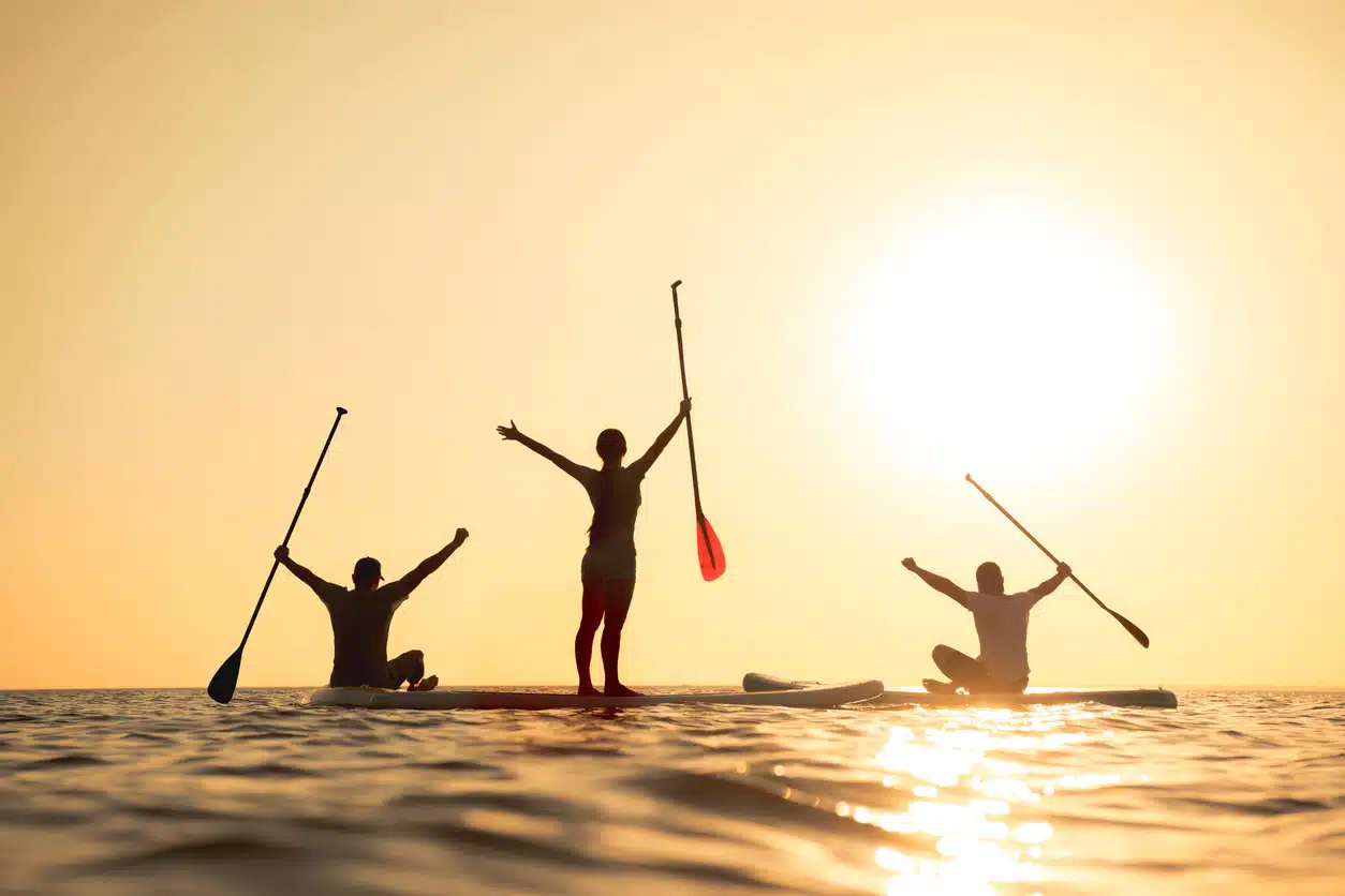 Happy surfers stands on sup boards with raised arms and looks at sunset