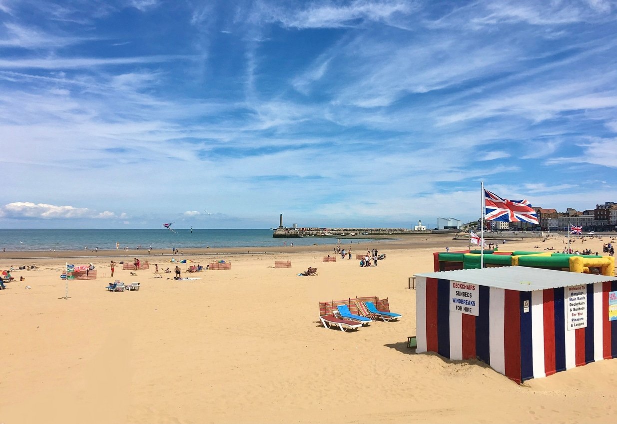 Margate, the great British seaside town. Golden sands and blue skies, people enjoying the beach and a hut with a British flag in the foreground