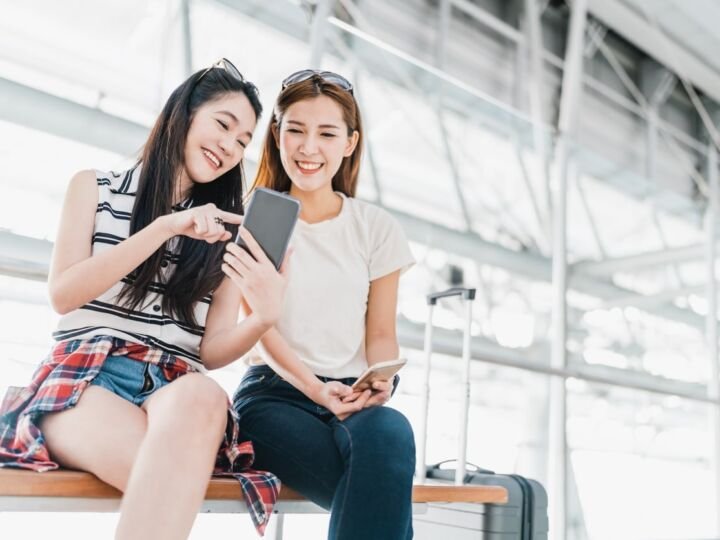 Two happy Asian girls using smartphone checking flight or online check-in at airport together, with luggage. Air travel, summer holiday, or mobile phone application technology concept