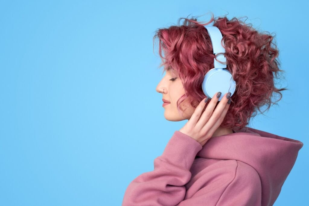 Profile view portrait of dreamy young pink hair girl wearing in casual clothes, enjoying sound and listening music in headphones standing against blue background with copy space