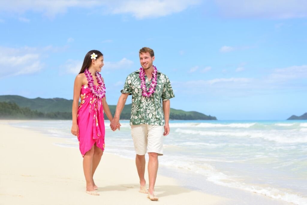 Happy couple on Hawaii vacation walking on beach with Hawaiian leis and Aloha clothing. Caucasian man wearing typical Hawaiian shirt and Asian woman girlfriend in pink sarong fabric sundress.