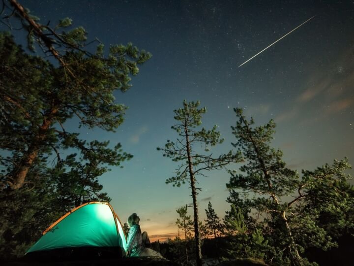 Man looking up at night starry sky with falling star and his tent lit up below