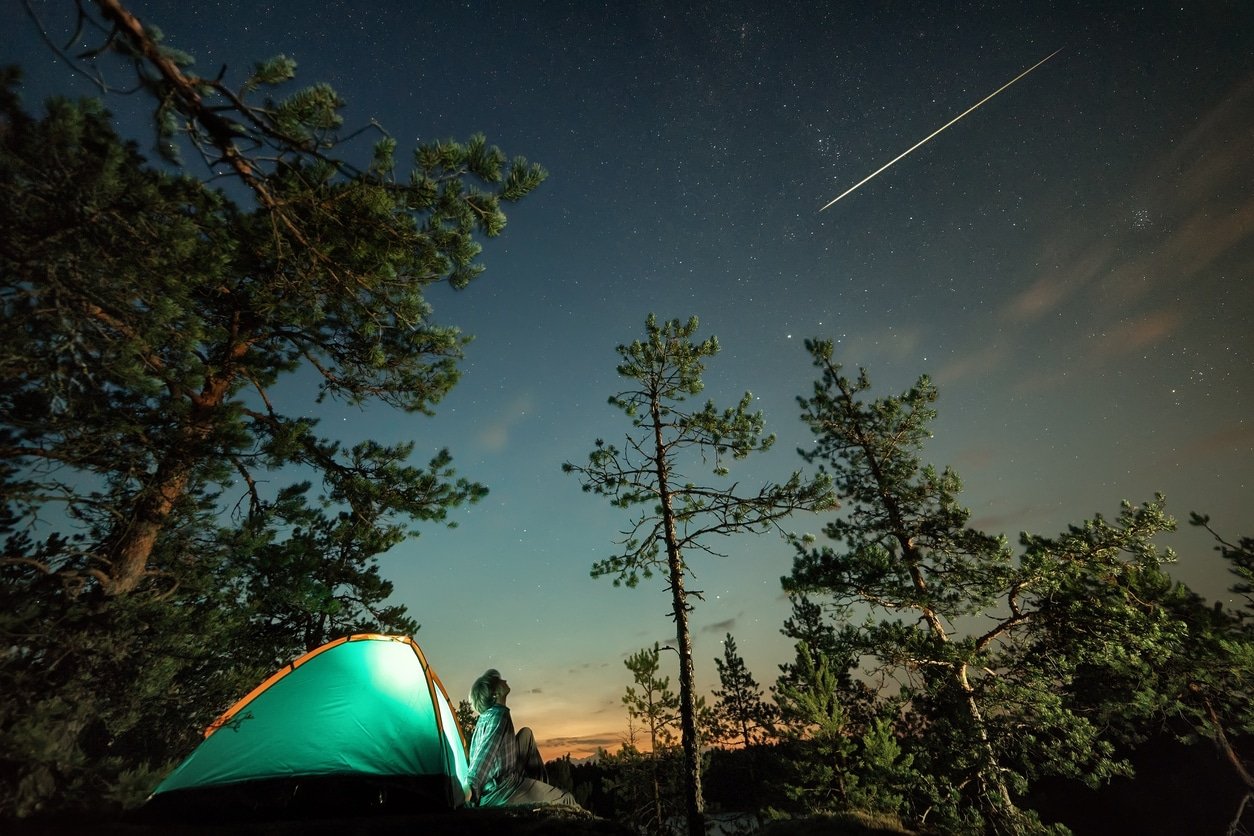 Man looking up at night starry sky with falling star and his tent lit up below