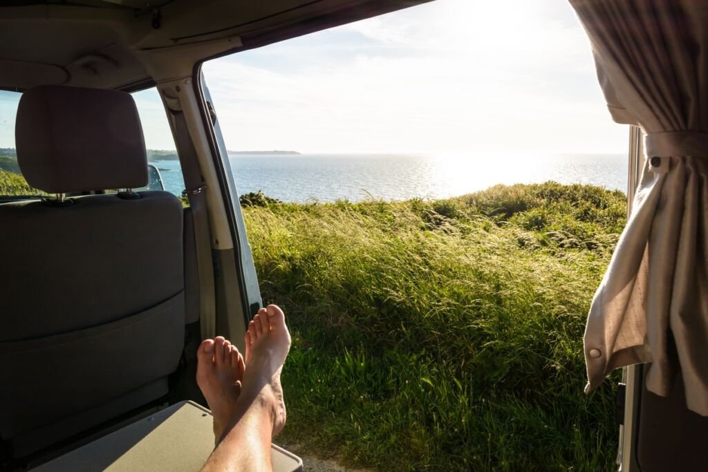 First-person view of a barefoot man relaxing inside a camper van and enjoying the view over the sea at sunset through the open sliding door with wild grasses in the foreground.
