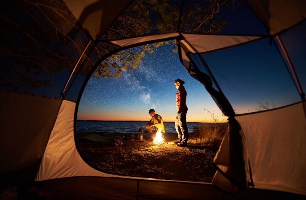 Young couple man and woman having rest at tent and burning campfire on sea shore near forest