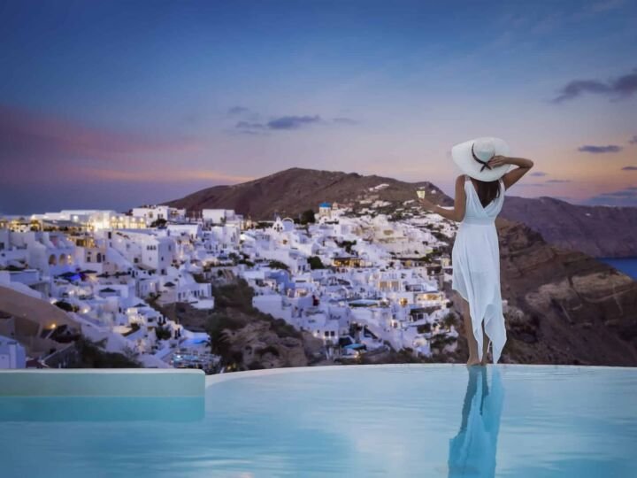 A woman in a white dress stands by the swimming pool and enjoys the view over the illuminated village of Oia, Santorini island, Greece, during summer sunset time