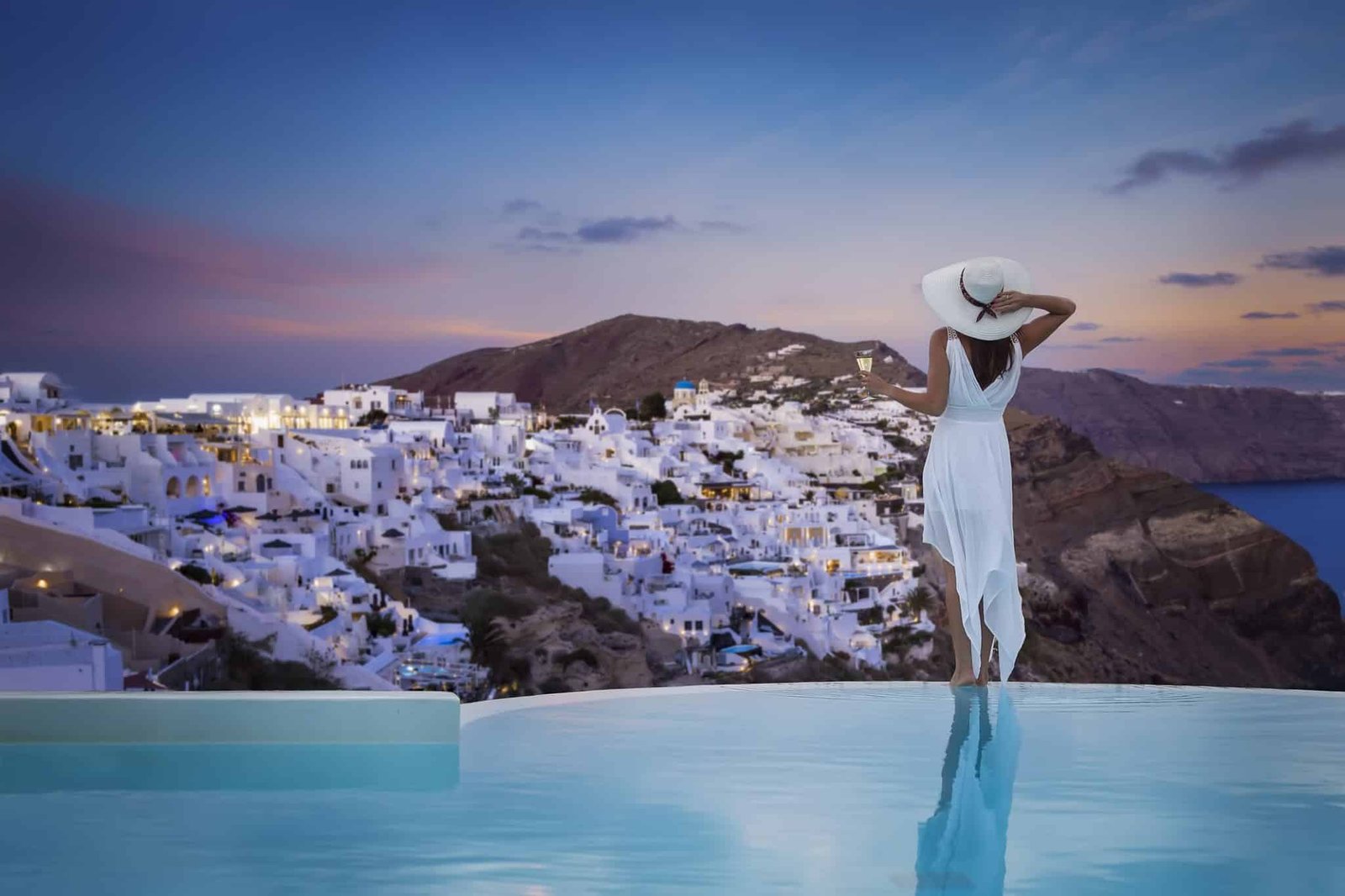 A woman in a white dress stands by the swimming pool and enjoys the view over the illuminated village of Oia, Santorini island, Greece, during summer sunset time