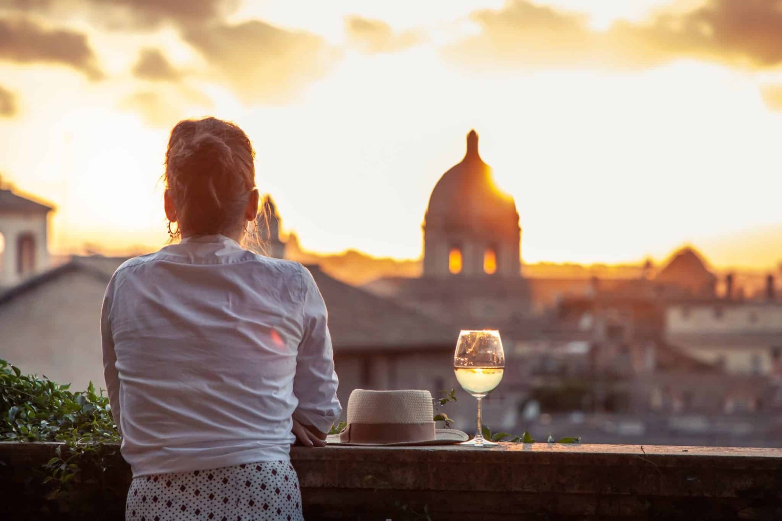 Young woman tourist fashion white dress with glass of white wine in front of panoramic view of Rome cityscape from campidoglio terrace at sunset. Landmarks, domes of Rome, Italy.