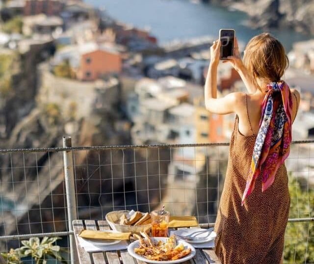 Woman takes photo of beautiful coastline in Vernazza village, traveling in famous Cinque Terre region in northwestern Italy. Girl sitting at outdoor restaurant with mediterranean food