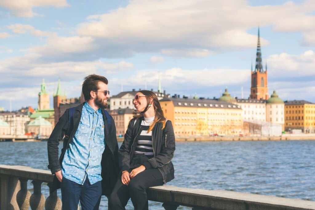 Young hipster couple in Stockholm. They are looking each other and flirting with sea and old town on background. Both are wearing sunglasses and a black jacket. Love and valentines day concepts.