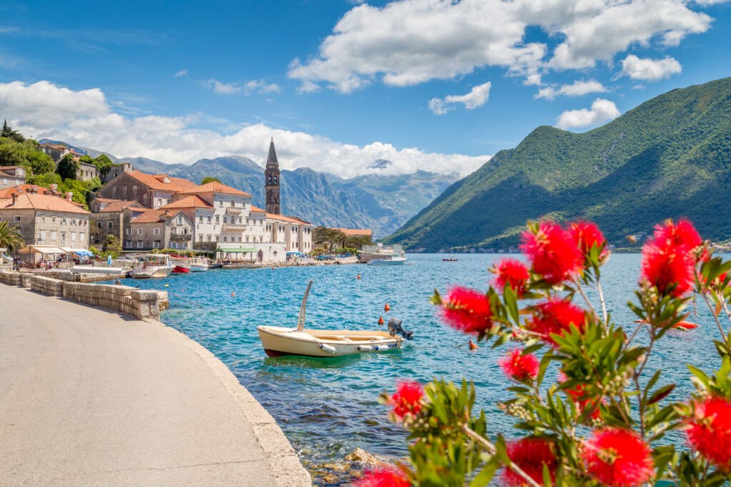 On a sun-drenched day in Perast, the Bay of Kotor shimmers beneath a vast blue sky, as the town's ancient buildings and verdant islets create a postcard-perfect coastal idyll.