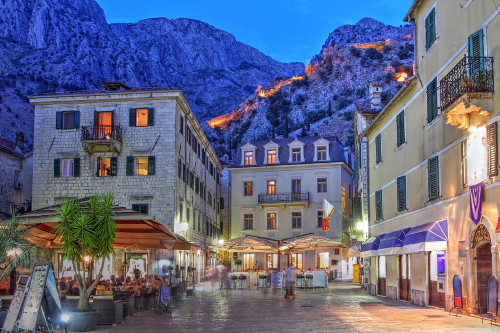 As twilight descends, Montenegro's ancient city walls aglow against the mountain backdrop create a mesmerizing silhouette, blending history with the majestic natural grandeur of the rugged landscapes.