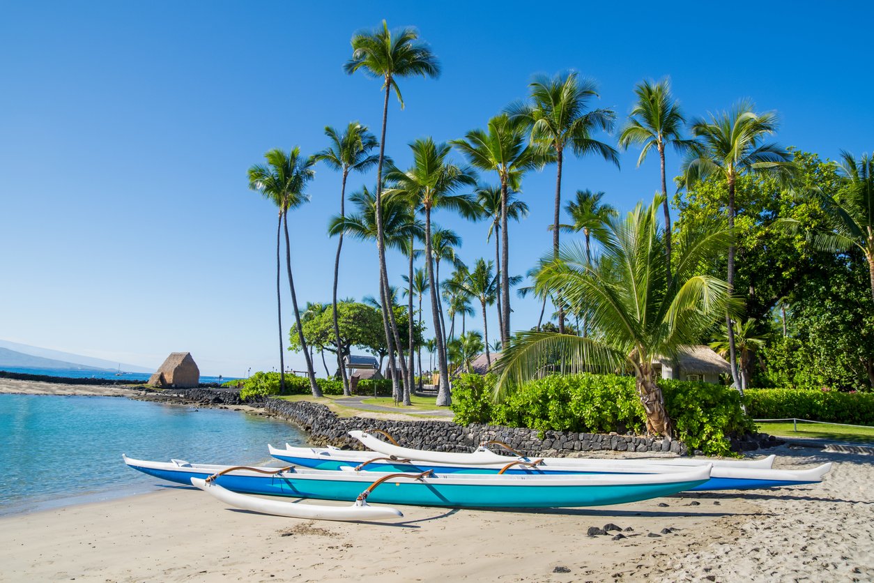 A Hawaiian outrigger canoe, vibrant against Kamakahonu Beach's golden sands. The Big Island's sun kisses the watercraft, echoing the spirit of ancient Polynesian voyages in modern coastal bliss.