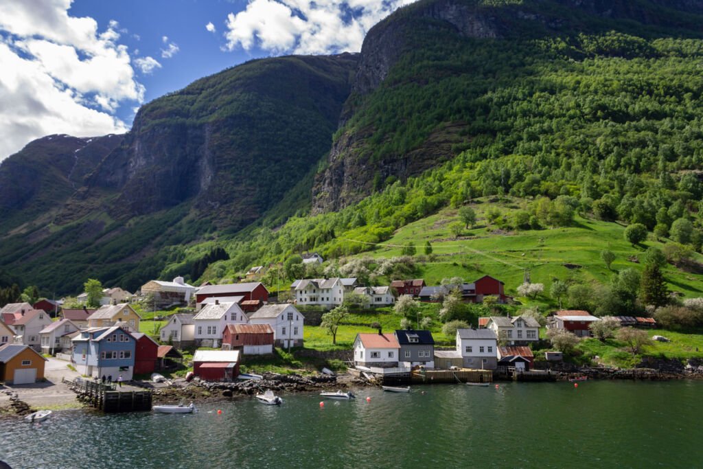 Houses grace the Narrow Fjord's edge in Flam village, Norway. By the lake, they stand, reflections rippling beneath snow-capped peaks, capturing the tranquil beauty of this coastal haven.