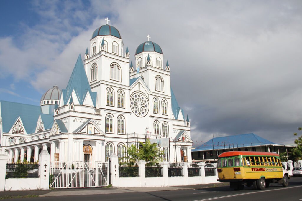 In the heart of Apia, Samoa, the Immaculate Conception of Mary Cathedral stands adorned. A local bus weaves past, adding life to the serene scene of this central city treasure.