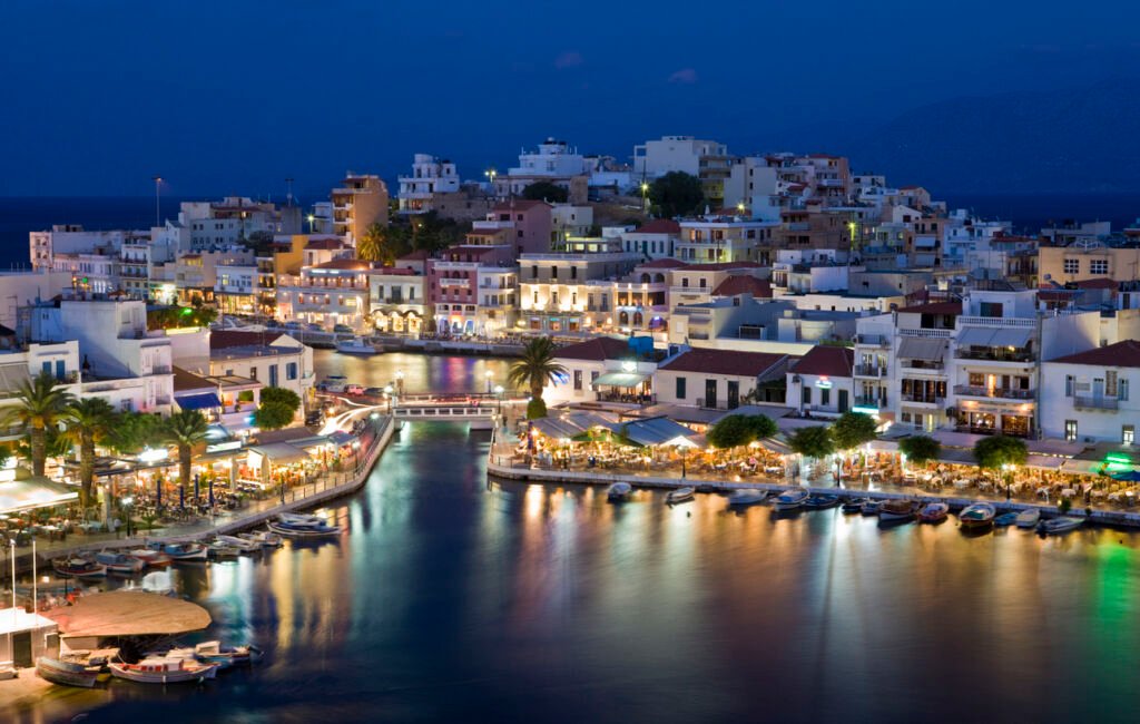 Agios Nikolaos in Crete shimmers enchantingly at night, its harbor a tapestry of twinkling lights reflecting on the calm waters, while the moon casts a gentle glow over the picturesque town.