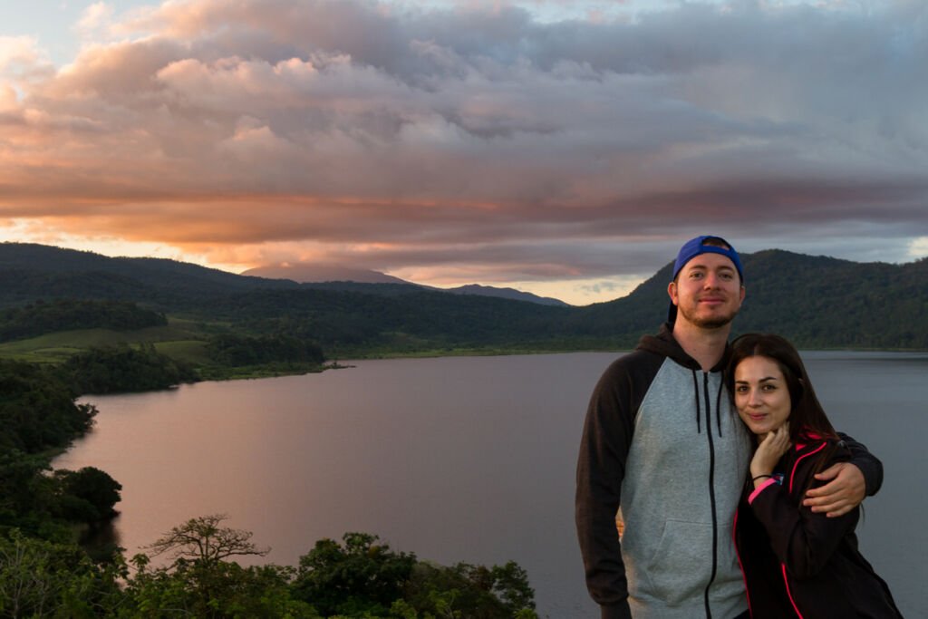 At Cote Lake, Costa Rica, a young couple embraces as the sunset paints the sky. Warm hues reflect on clouds, creating a romantic canvas above, capturing love in nature's glow.