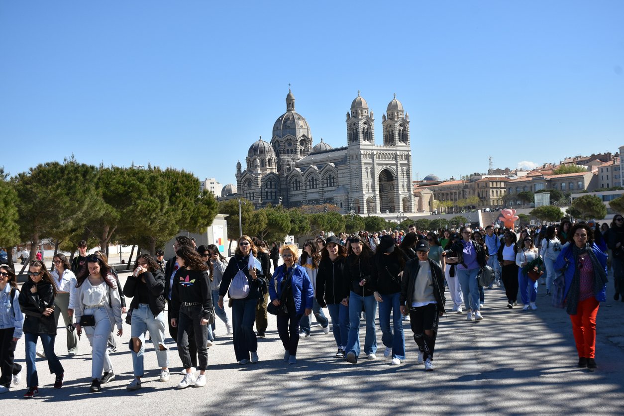 In Marseille, Europe, the Cathedral de la Major stands grand against the spring sky. People stroll beneath blooming trees, harmonizing the historic architecture with vibrant life and movement.