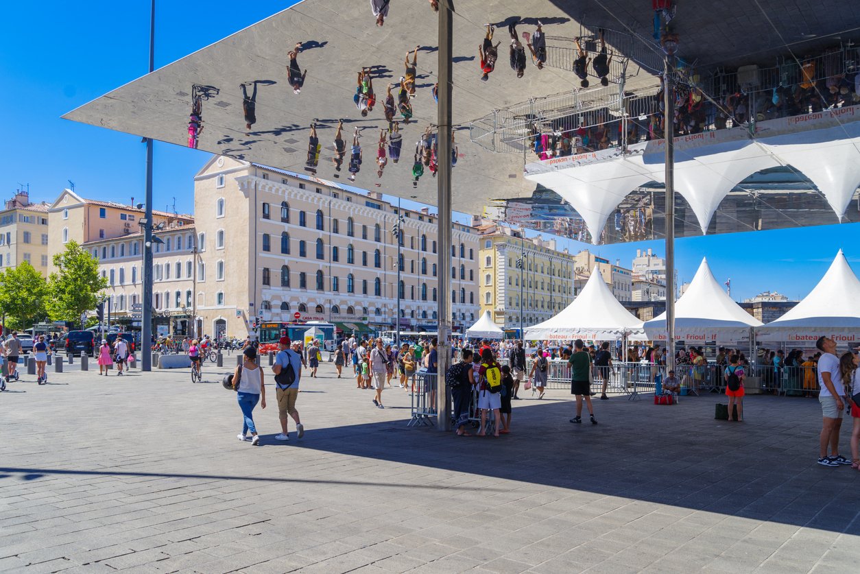 Marseille's Old Harbor buzzes with life. Beneath a mirrored canopy, people gather in the ancient natural harbor turned vibrant, pedestrian-friendly hub, echoing centuries of maritime history.