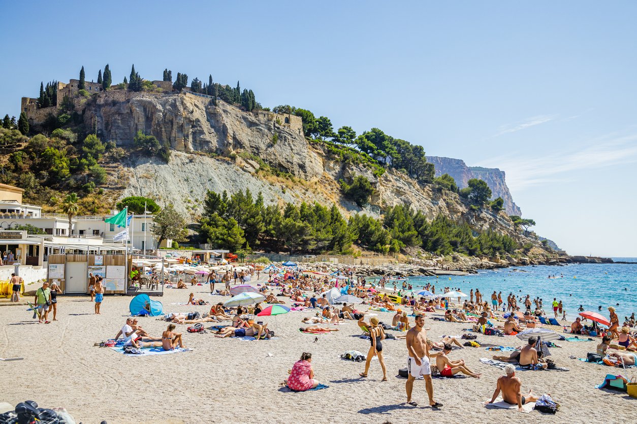 Plage de la Grande Mer in Cassis, Provence, hums with vibrant energy. Sun-soaked sands host a mosaic of umbrellas and joyful vacationers, as azure waves embrace the lively summer scene.
