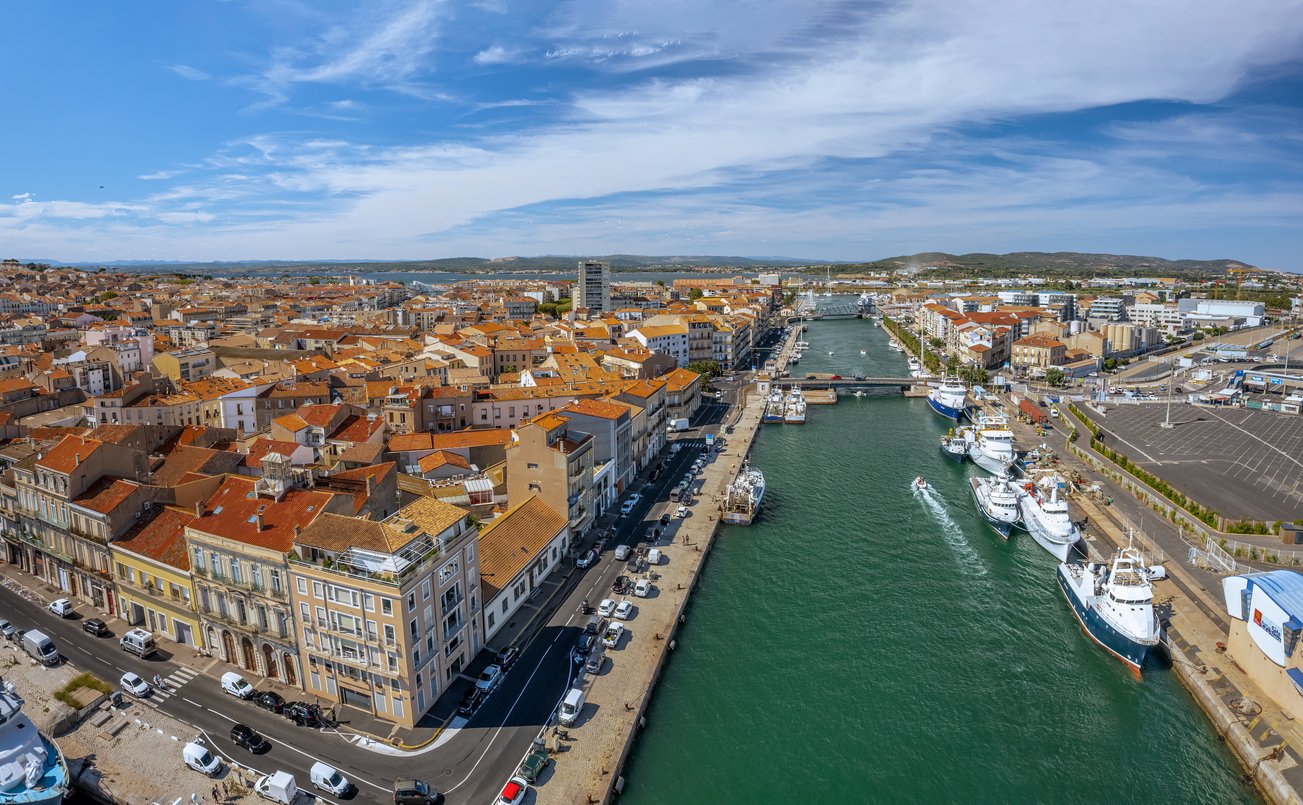 Sète's Royal Canal unfolds like a liquid ribbon from a drone's perspective. Quaint boats dot the water, flanked by colorful buildings, creating a picturesque tableau in Occitanie, France.