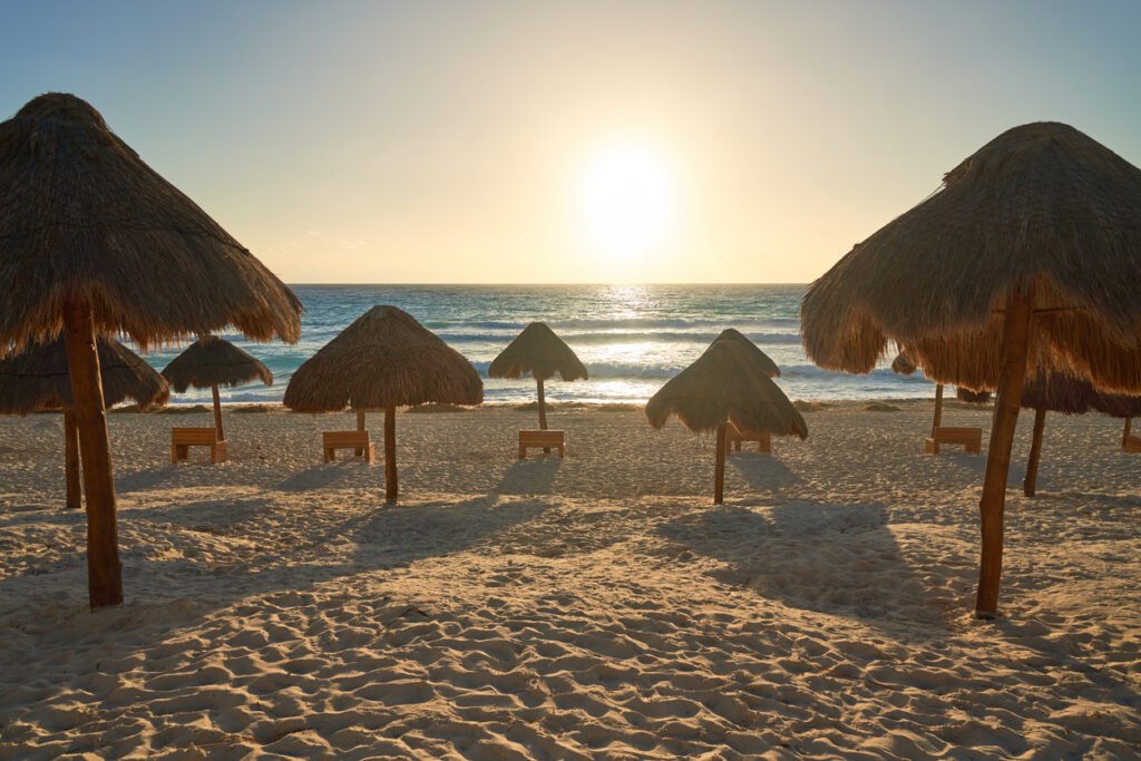Rustic umbrellas grace the shores of Delfines Beach in Cancun, Mexico, as the sun paints the sky with hues of sunrise or sunset. A tranquil scene blending nature's beauty with coastal charm.