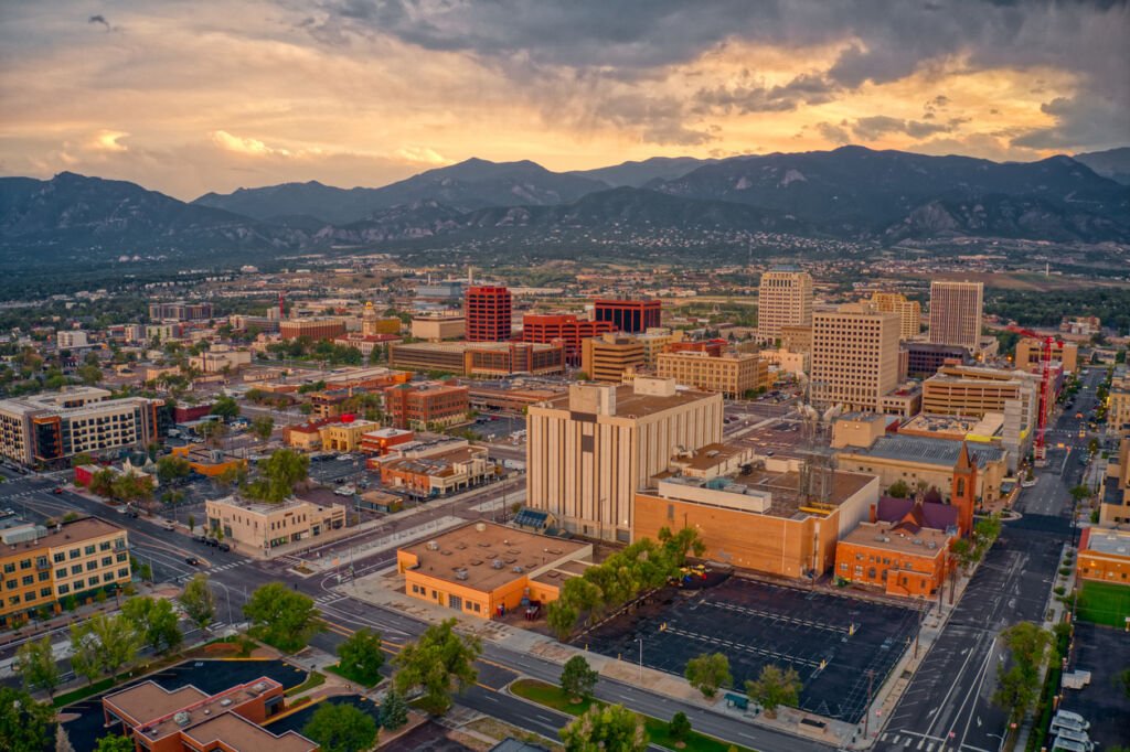 Bathed in the soft hues of dusk, an aerial panorama unfolds over Colorado Springs, revealing a cityscape aglow with city lights and nestled against the majestic silhouette of the surrounding mountains.