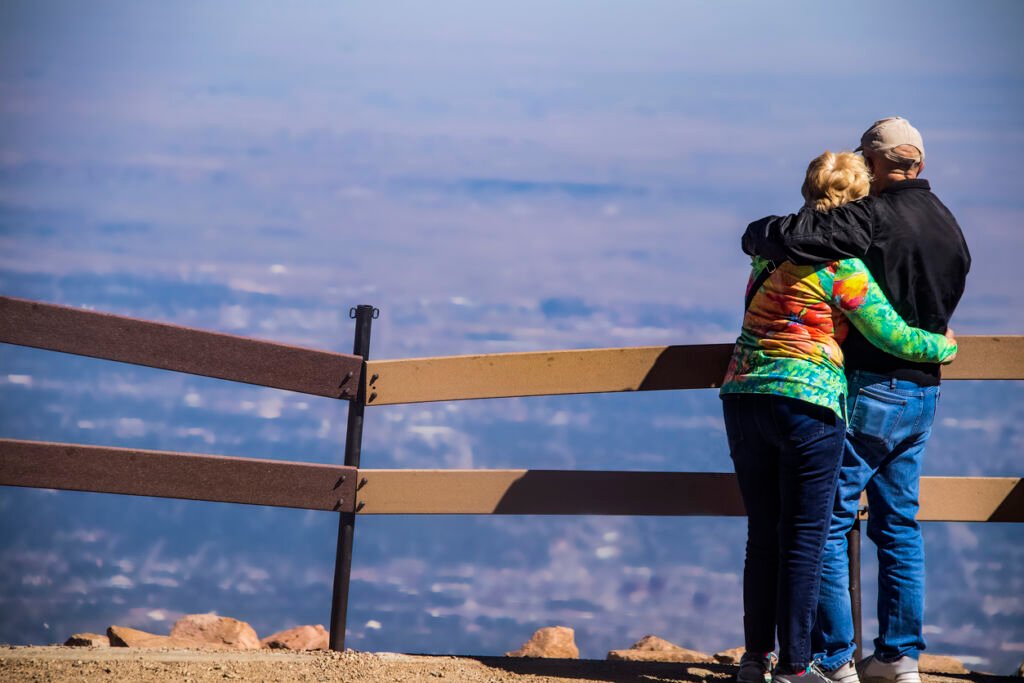 In the Colorado mountains, USA, an older couple stands united, arms around each other, atop a summit. They share a moment of tranquility, gazing down at a breathtaking panoramic view.