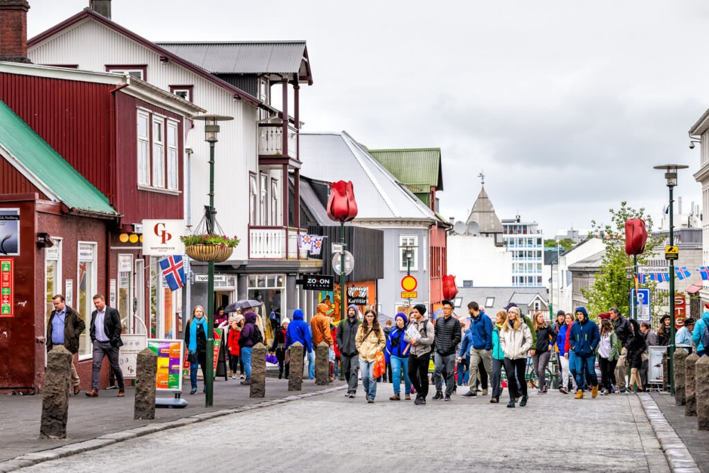 Reykjavik, Iceland: A bustling summer scene unfolds in the downtown center. Tourists fill the sidewalks, exploring stores and restaurants adorned with vibrant signs, creating a lively atmosphere.