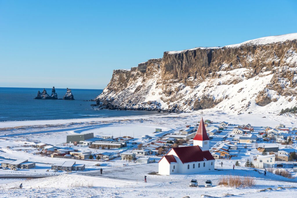 In the winter wonderland of Iceland, Vik I Myrdal church crowns the hill, offering a breathtaking panoramic view of the picturesque village. The snow-covered landscape enhances the beauty of this enchanting scene.