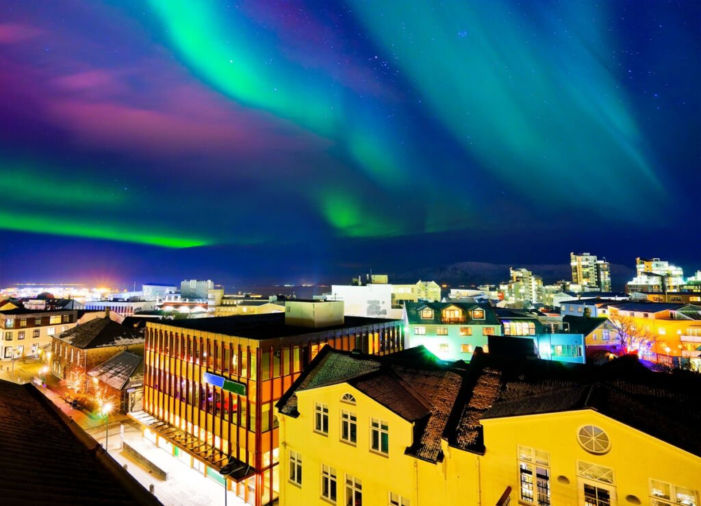 The city center of Reykjavik, Iceland, transforms into a magical scene as the northern lights dance across the night sky. Vibrant hues of green and purple illuminate the urban landscape, creating a celestial display above the city lights.