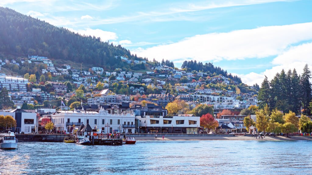 Queenstown's cityscape unfolds along its enchanting harbor, adorned with colorful buildings and boats. The view extends across the water, revealing a charming marina and picturesque shoreline, capturing the essence of this lakeside gem.