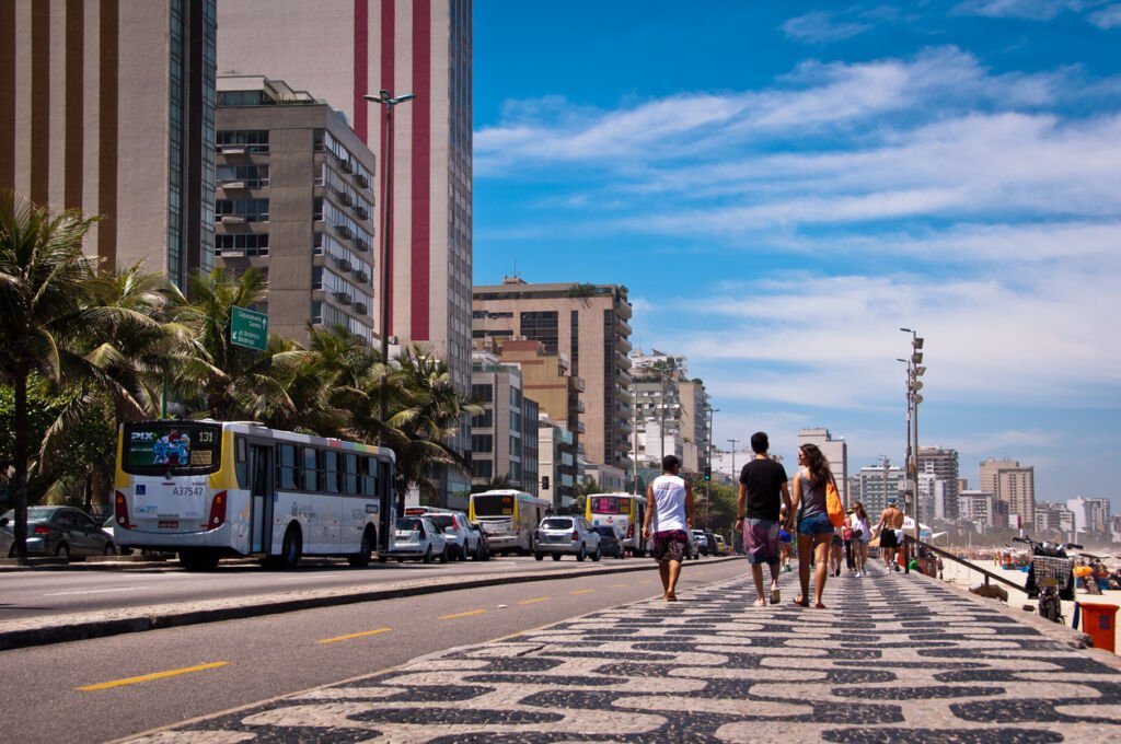 In Rio de Janeiro, Brazil, people leisurely stroll along the sidewalk of Avenida Vieira Souto in Ipanema. The vibrant atmosphere and scenic views contribute to the lively urban experience.
