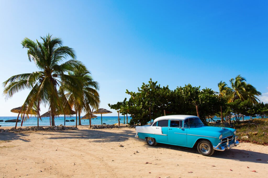 An iconic vintage car, bathed in golden sunlight, rests serenely on the sandy shores of a tranquil Cuban beach, embodying timeless charm and Caribbean allure.
