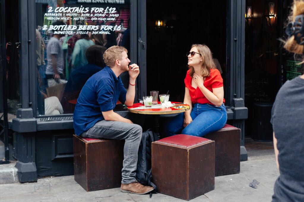 On Monmouth Street, London, young people gather outside a cozy bar or coffee shop, savoring drinks and lively conversation amidst the vibrant urban atmosphere.
