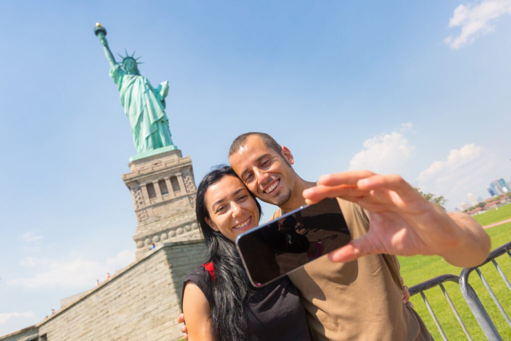 A couple captures a cherished moment, smiling as they take a selfie with the majestic Statue of Liberty standing tall in the background, a timeless symbol of freedom and unity.