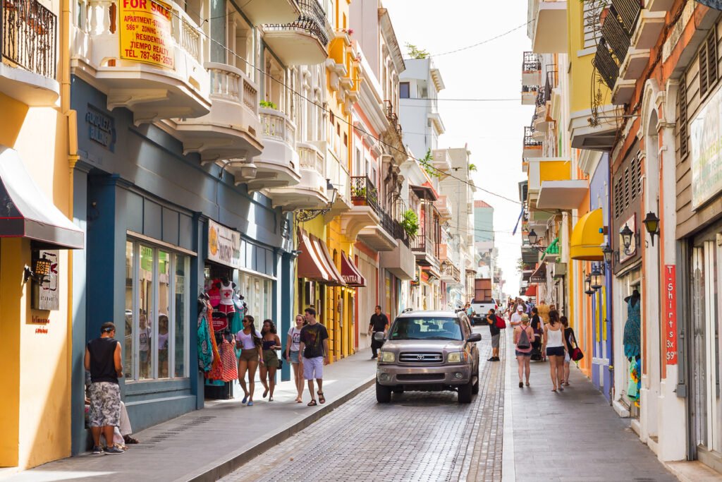 In San Juan, Puerto Rico, the main street bustles with life as people weave through vibrant shops, their laughter and chatter echoing the lively spirit of the city.